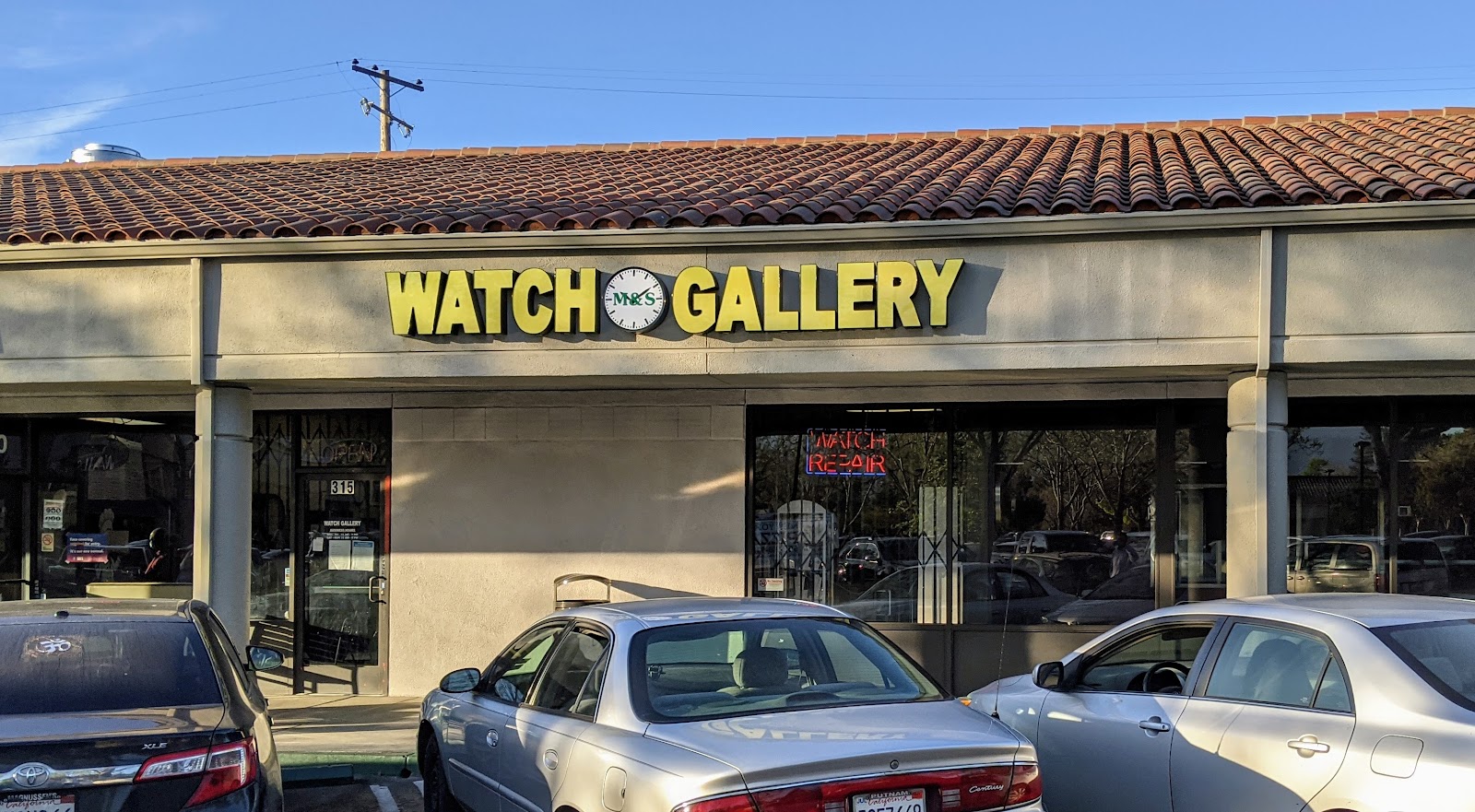 M & S Watch Gallery storefront in Mountain View — bold yellow WATCH GALLERY sign on a dark backdrop with a clock-face M&S logo, at golden hour.